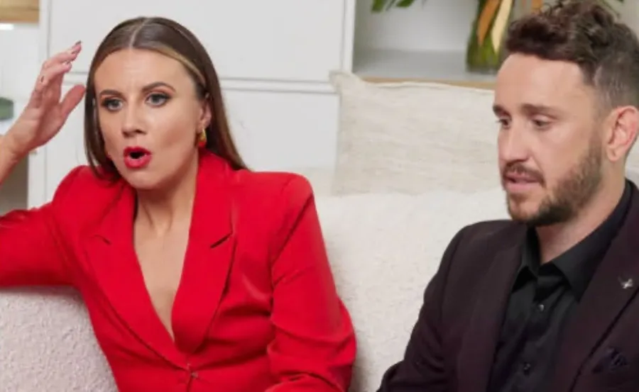 A woman in a red outfit and a man in black sit on a couch, both appearing surprised or engaged in conversation.