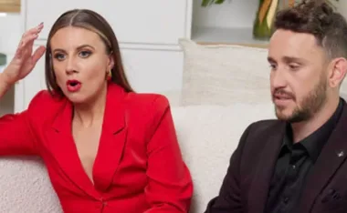 A woman in a red outfit and a man in black sit on a couch, both appearing surprised or engaged in conversation.