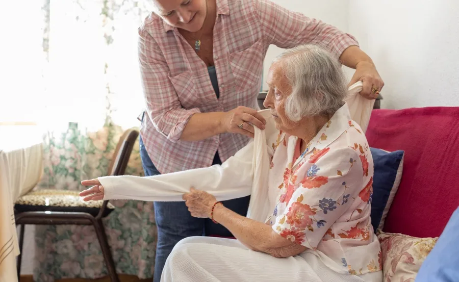 A caregiver helps an elderly woman in a floral blouse put on a sweater while seated on a sofa.