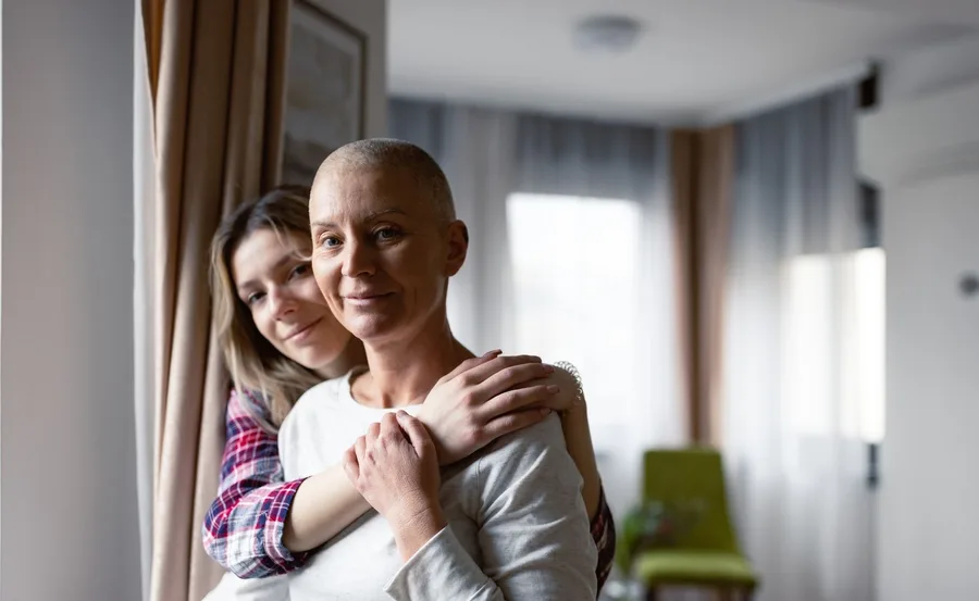 Mother with cancer embraced by daughter, standing in a cozy room, highlighting warmth and support.
