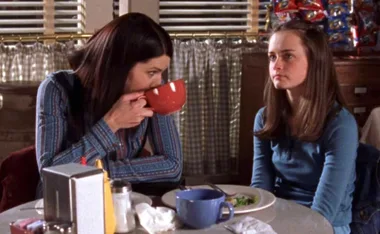 Two women sitting at a diner table, one sips from a large red mug, the other looks ahead with a neutral expression.
