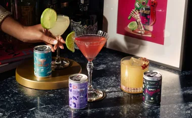 Cans of Curatif cocktails with garnished drinks on a bar table, featuring Gimlet, Cosmopolitan, and Spicy Margarita.