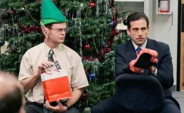 Two men sitting by a Christmas tree; one in an elf hat holds a gift bag, the other in a suit holds oven mitts.