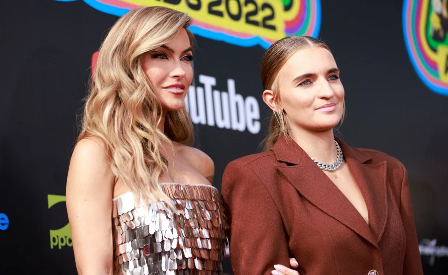 Two women posing on the red carpet; one in a silver dress and the other in a brown blazer.