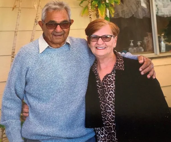 Elderly couple smiling, standing together outside a house with plants in the background.