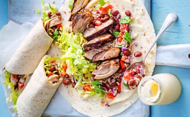 Grilled chicken burritos with lettuce, beans, rice, and sauce on tortillas, with two wrapped ones on the side.