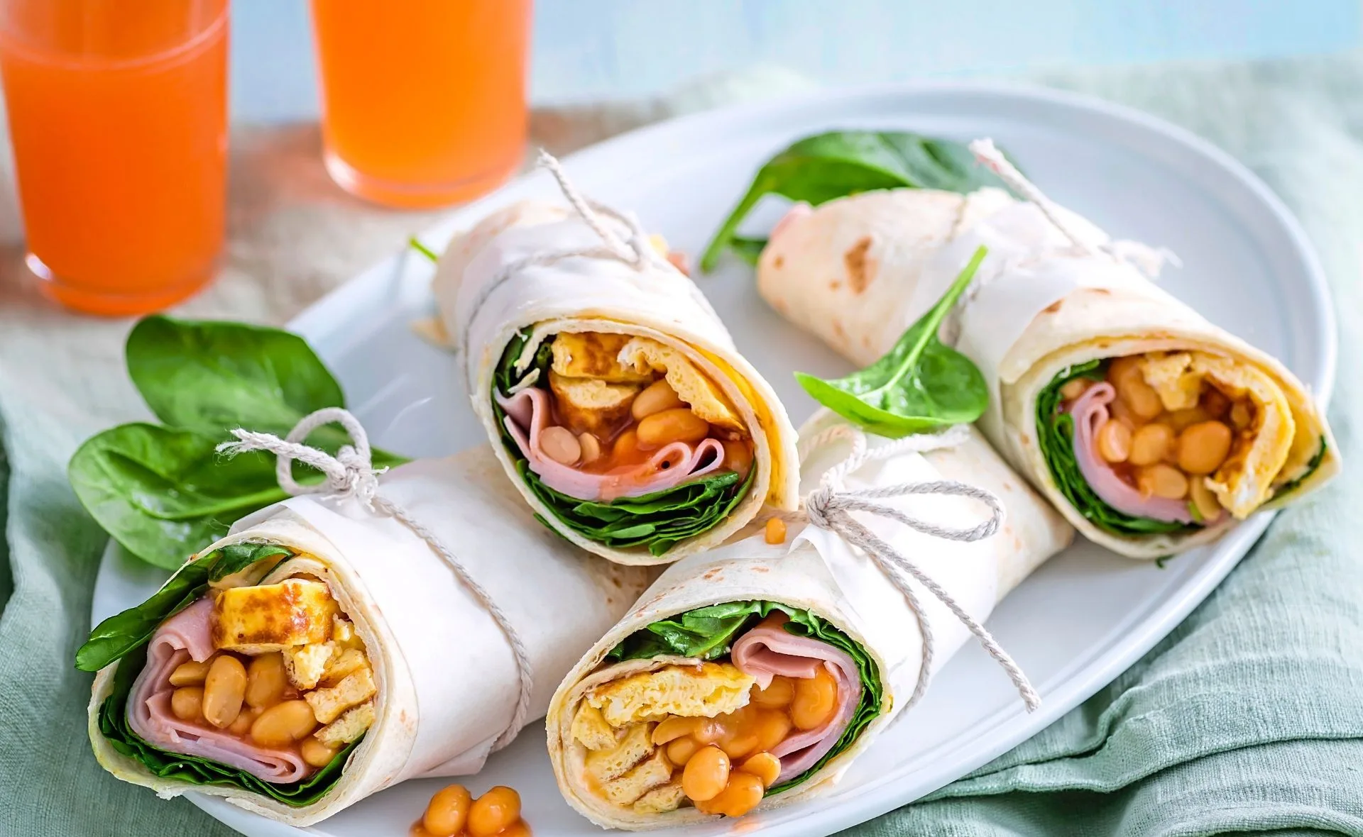 Breakfast burritos wrapped with twine on a plate, containing eggs, beans, ham, and spinach, with two orange juices.