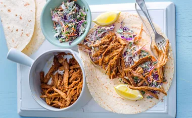 Pulled pork and coleslaw on a tortilla with lemon wedges, served on a white cutting board.