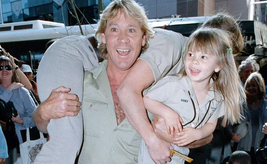 Steve Irwin joyfully carries Bindi Irwin on his arm, surrounded by a crowd, both smiling and wearing matching khaki outfits.