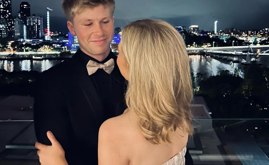 A couple embracing on a balcony at night, overlooking a lit-up city skyline and river.