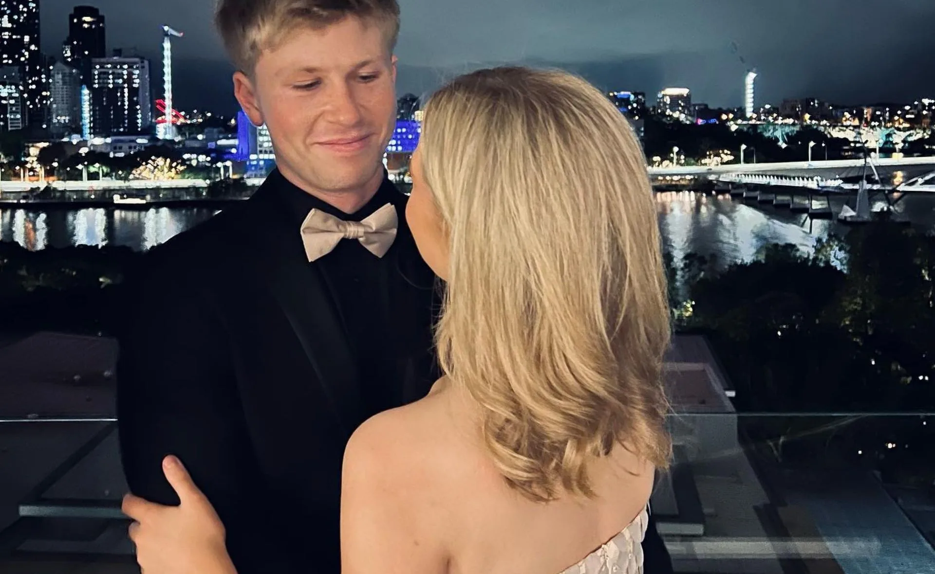 A couple embracing on a balcony at night, overlooking a lit-up city skyline and river.