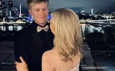 A couple embracing on a balcony at night, overlooking a lit-up city skyline and river.