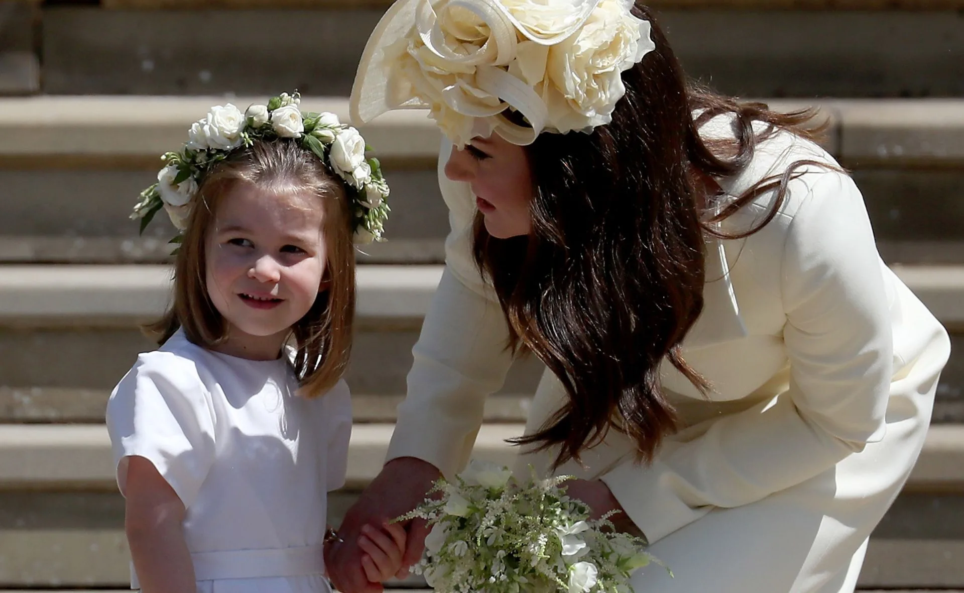 Woman in a cream outfit and hat talking to a young girl wearing a white dress and flower crown.