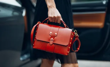 Person holding a stylish red handbag with gold studs next to a car.