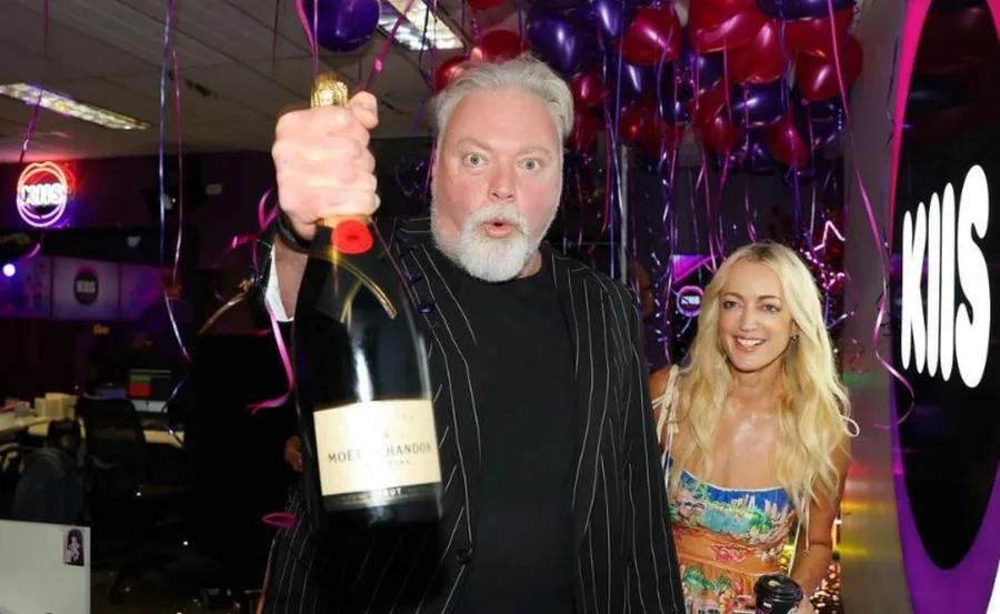 Man holding champagne with a surprised expression, woman smiling, in a festive room with KIIS logo and balloons.