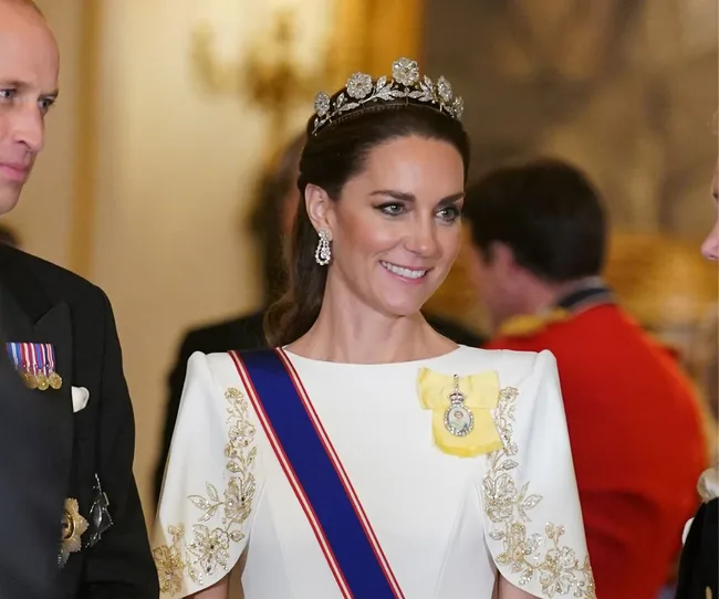 Princess wearing a jeweled tiara and white dress with gold embroidery in a formal setting.