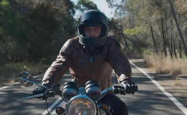 Remi rides a motorcycle on an empty rural road, wearing a leather jacket and helmet.