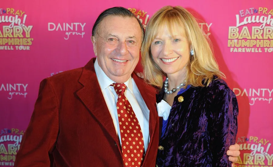 Two people smiling at a Barry Humphries farewell event, in front of a pink backdrop.