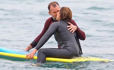 A couple in wetsuits embrace on a surfboard in the water, surrounded by gentle waves.