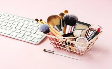 A shopping basket filled with makeup products and brushes beside a white keyboard on a pink background.