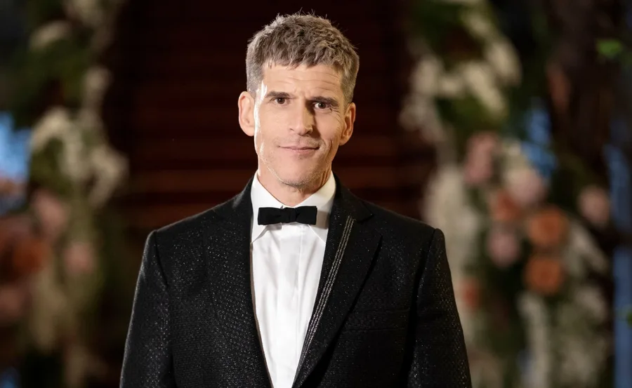 A man in a black tuxedo with a bow tie standing in front of a blurred floral and staircase background.