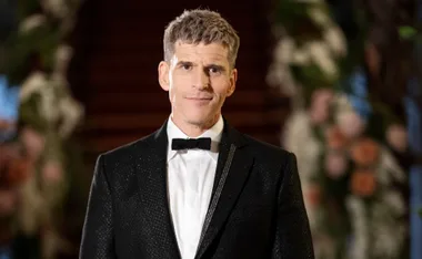 A man in a black tuxedo with a bow tie standing in front of a blurred floral and staircase background.