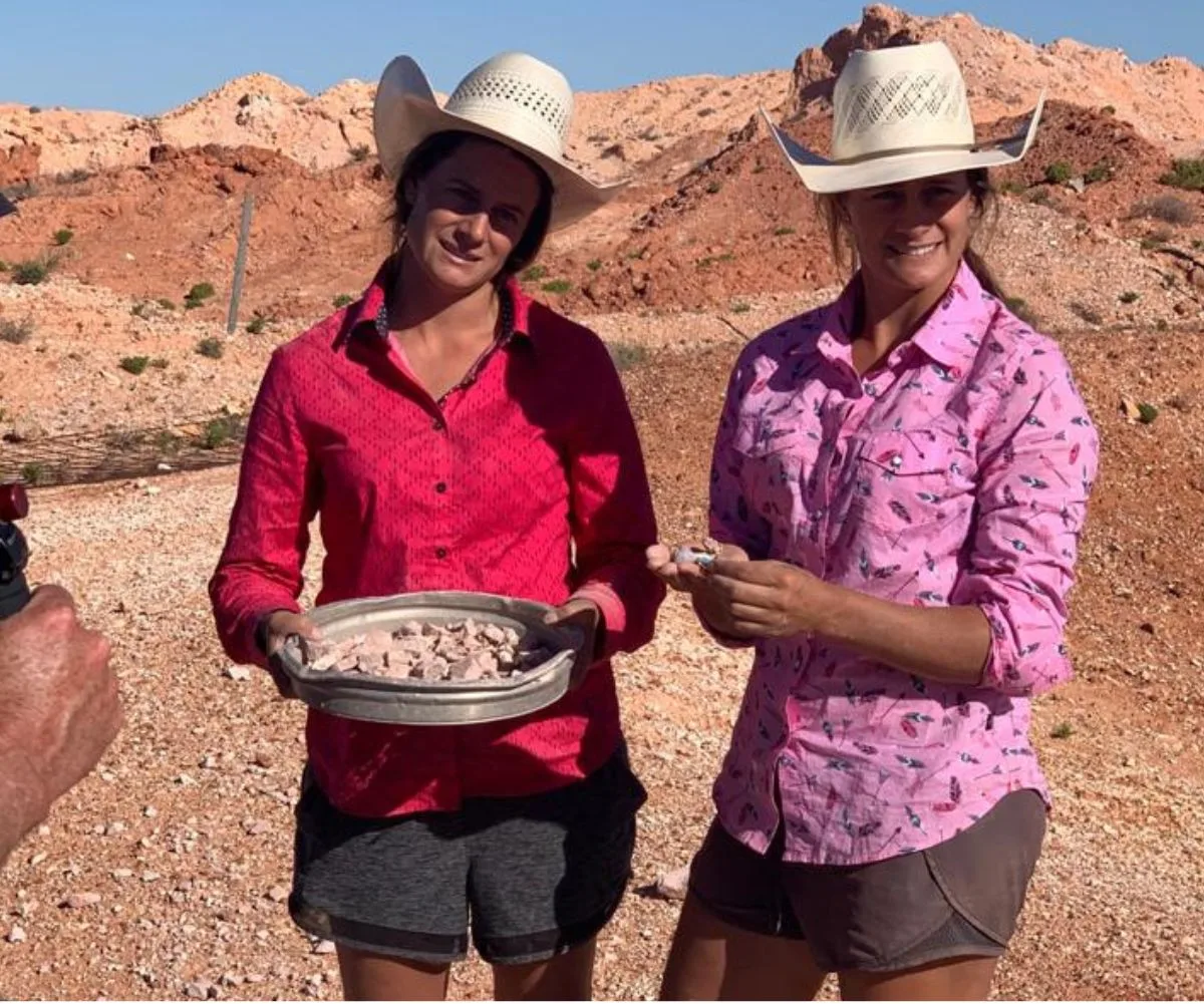 Two people in cowboy hats and colorful shirts holding a tray of rocks in a rocky desert landscape.