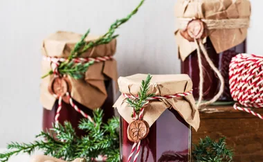 Jars of jam wrapped in brown paper with twine, decorated with evergreen sprigs and wax seals, beside red-white string.