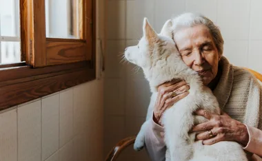 Elderly woman hugging a fluffy white dog near a wooden window, expressing warmth and affection.