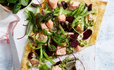 Salmon and beetroot tart with asparagus, peas, and leafy greens on a white background, partially cut with a knife.