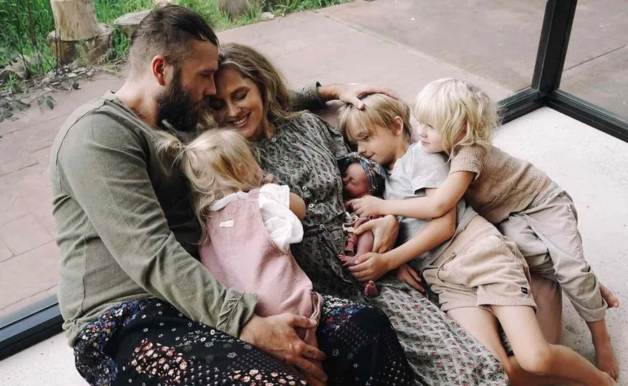 Family sitting together on floor, smiling and cuddling, with young children embracing a newborn.