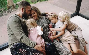 Family sitting together on floor, smiling and cuddling, with young children embracing a newborn.