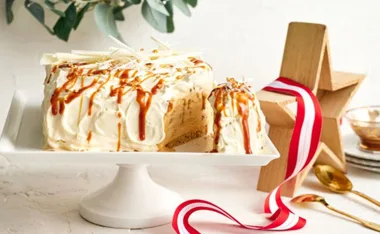 Creamy banana bread ice cream cake topped with caramel drizzle and white chocolate shavings on a white pedestal.