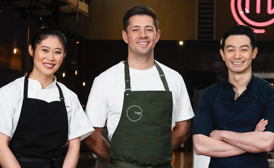 Three chefs smiling, wearing aprons in a kitchen setting with a MasterChef logo in the background.