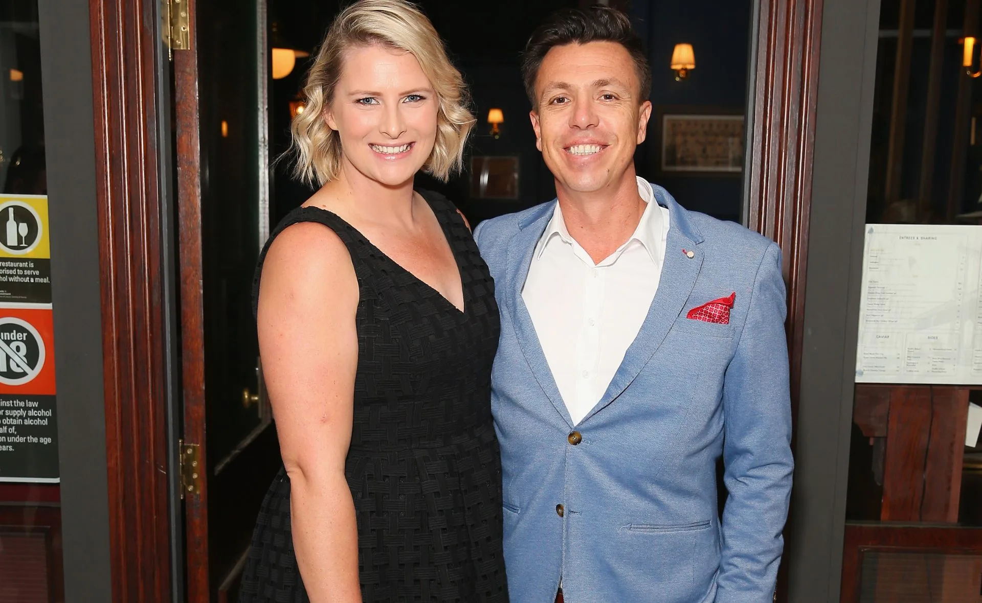 A smiling couple in formal attire, woman in black dress and man in blue blazer, standing together indoors.