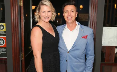 A smiling couple in formal attire, woman in black dress and man in blue blazer, standing together indoors.
