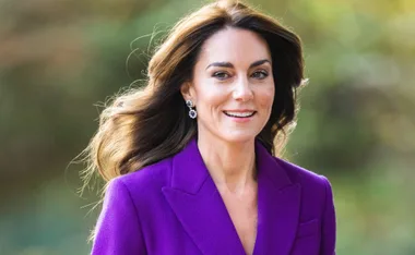 A woman in a purple coat, smiling outdoors with flowing hair and elegant earrings.