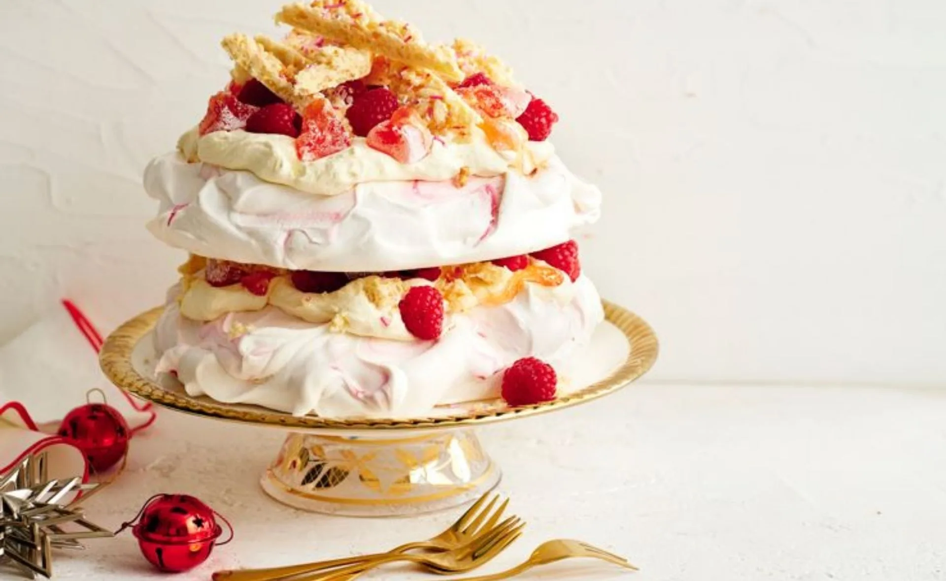 Elegant white pavlova topped with raspberries and almond slivers on a gold plate; festive decorations nearby.