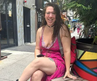A woman in a pink dress sits smiling on a city street bench, holding a phone.