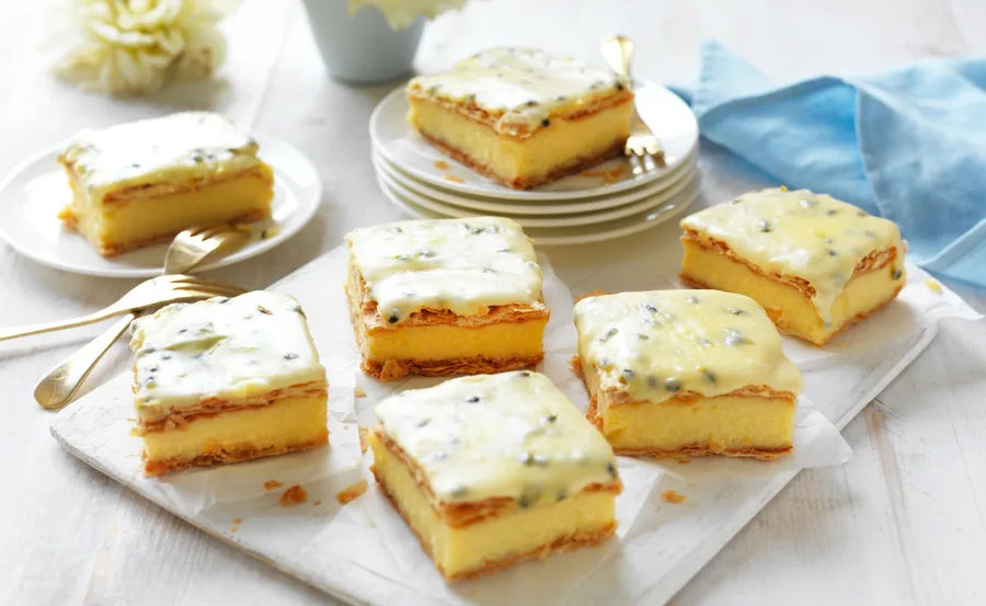 Vanilla slices with passionfruit icing on a white board, golden cutlery, blue napkin, and plates in the background.