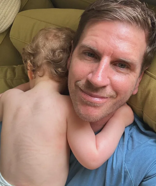 Man lying on a couch, smiling at the camera, with a baby sleeping on his chest.