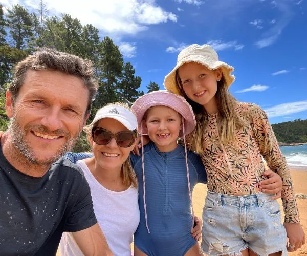 Family of four smiling at the beach with trees and ocean in the background.