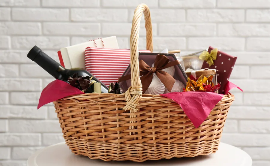 Christmas hamper with wrapped gifts, a wine bottle, and decorations in a wicker basket against a white brick wall.