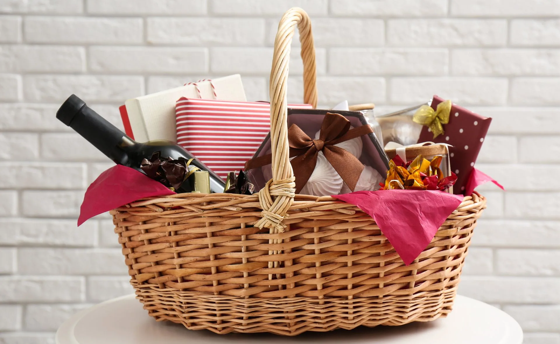 Christmas hamper with wrapped gifts, a wine bottle, and decorations in a wicker basket against a white brick wall.