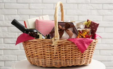 Christmas hamper with wrapped gifts, a wine bottle, and decorations in a wicker basket against a white brick wall.