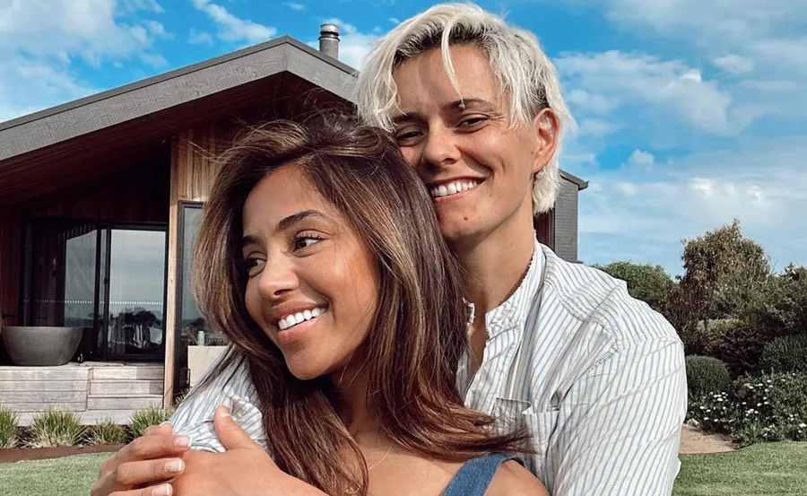 A smiling couple embraces in front of a modern wooden house on a sunny day.