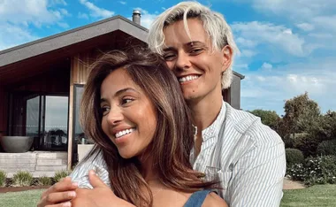 A smiling couple embraces in front of a modern wooden house on a sunny day.