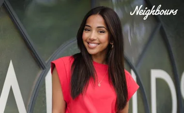 Young woman in a red top smiling, standing in front of a Neighbours TV show backdrop.