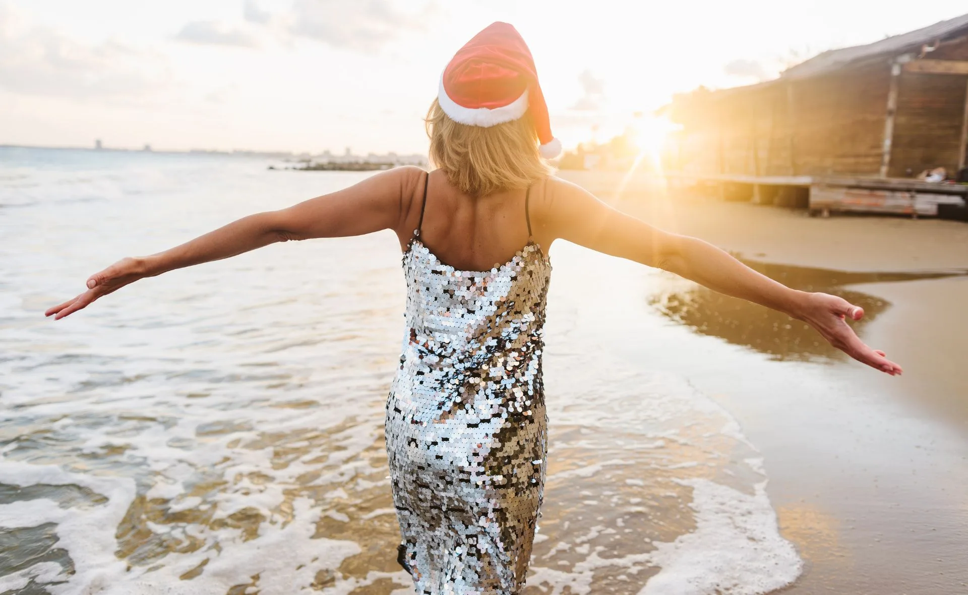 Person in a sequin dress and Santa hat spreading arms, walking on a sunny beach.