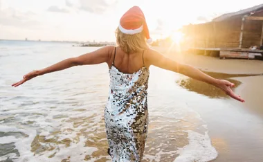 Person in a sequin dress and Santa hat spreading arms, walking on a sunny beach.